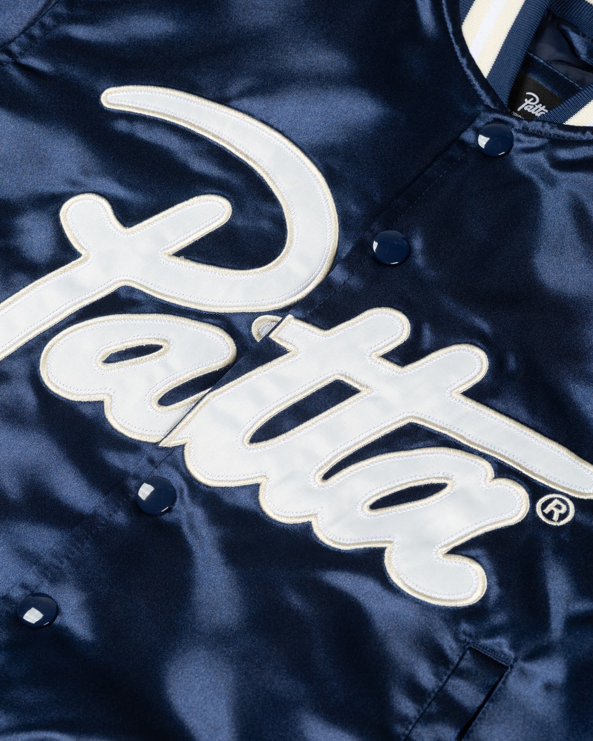 Detail of the white script logo appliqué on the Black Iris Patta kids satin bomber jacket.